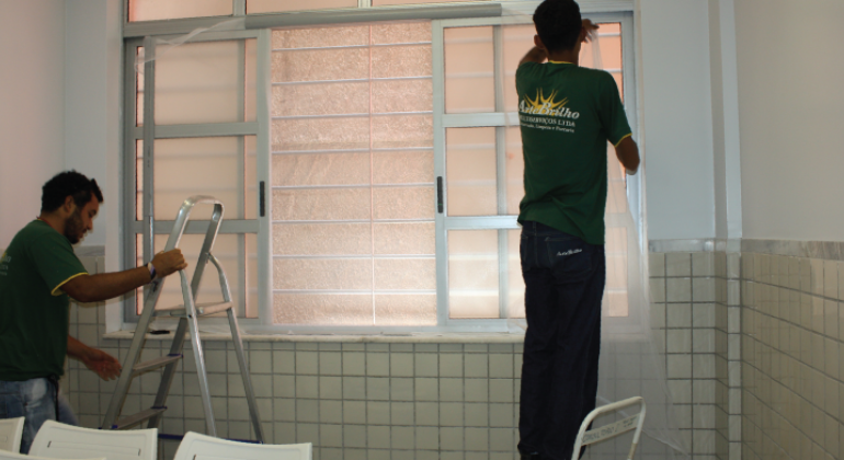Dois homens instalam tela em janela de centro de saúde.