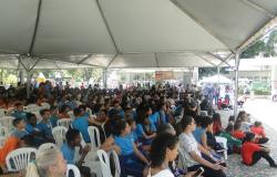 Mais de cinquenta pessoas sentadas em local coberto, assistindo a apresentações.