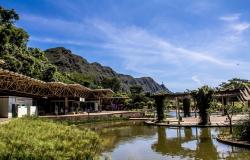 Parque das Mangabeiras com espelho d'água e grama à frente, área administrativa coberta com diversas salas e pessoas e caramanchão à direita. Ao fundo, Serra do Curral sob céu azul de dia. 