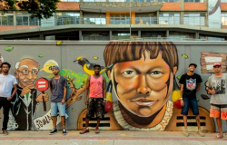 Quatro rapazes de bermuda e boné estão parados à frente de muro grafitado.