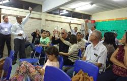 Cerca de cinco pessoas levantam a mão segurando cartões brancos em sala com mais de vinte pessoas, a maioria delas sentada.