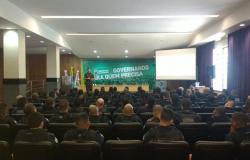 Grupo de cerca de 80 guardas estão sentados em um auditório, de costas para a foto. No palco, de frente, está um membro do exército brasileiro. Um banner escrito "Governando para quem precisa" está ao fundo.