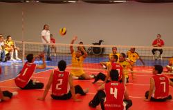 Jogo de vôlei com jogadores sentados: Na frente, time com uniforme vermelho e atrás, time com uniforme amarelo. A bola está no ar. 