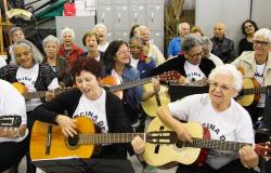 Cerca de treze idosos e idosas, todos sentados; cinco delas estão tocando violão.