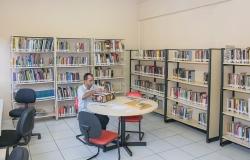 Homem sentado em mesa em biblioteca com várias estantes de livros. 