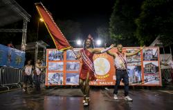 Passista de escola de samba é porta-bandeira, com vestido vermelho e faixa escrita "Madrinha do Leão". Ao lado, homem, representa o mestre-sala, e usa faixa com escrita "Padrinho do Leão". Os dois sambam na Avenida Afonso Pena à noite